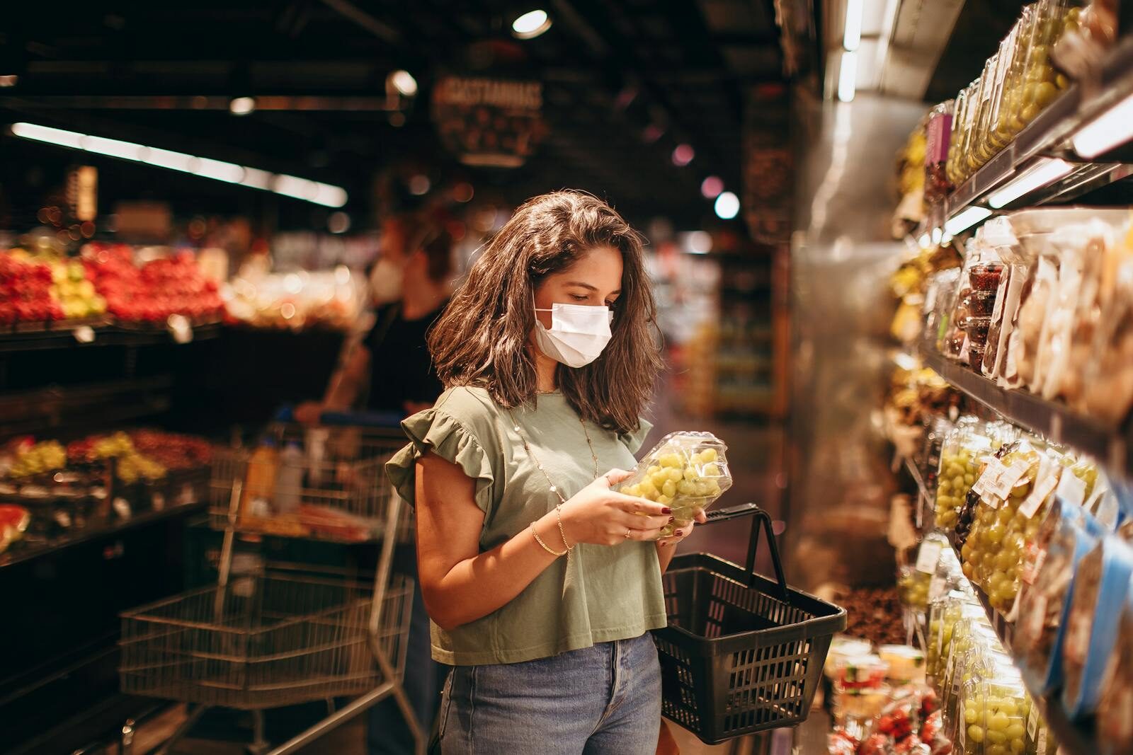 A woman wearing a mask inspects grapes in a supermarket aisle, holding a shopping basket.